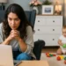 Žena sedí u laptopu s rukama složenýma a čte článek o zvýšení výživného na dítě, zatímco za ní si malé dítě hraje s barevnými hračkami na podlaze v útulném interiéru.